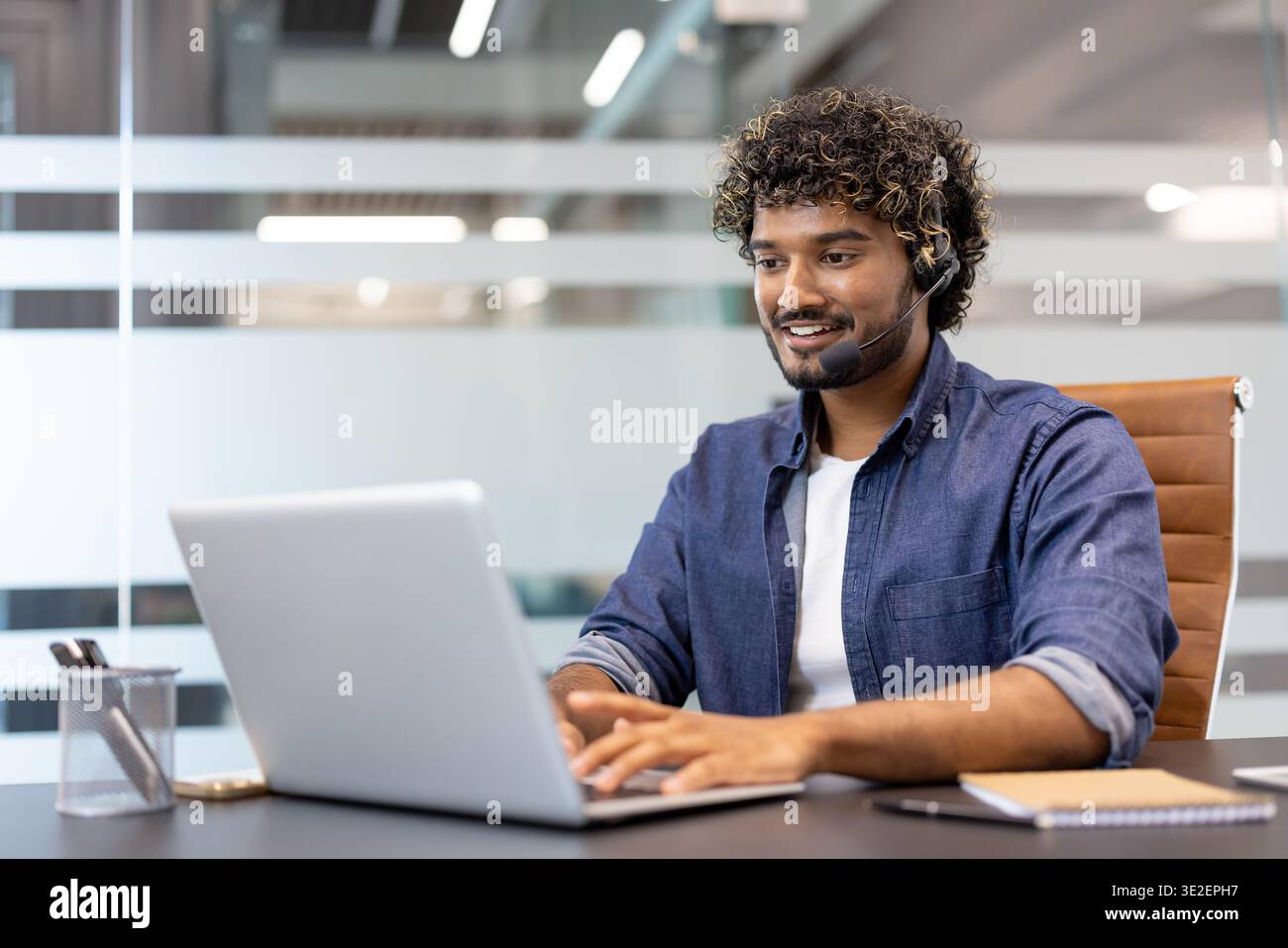 Junger indischer Mann im Headset, der im Büro am Laptop arbeitet. Stockfoto