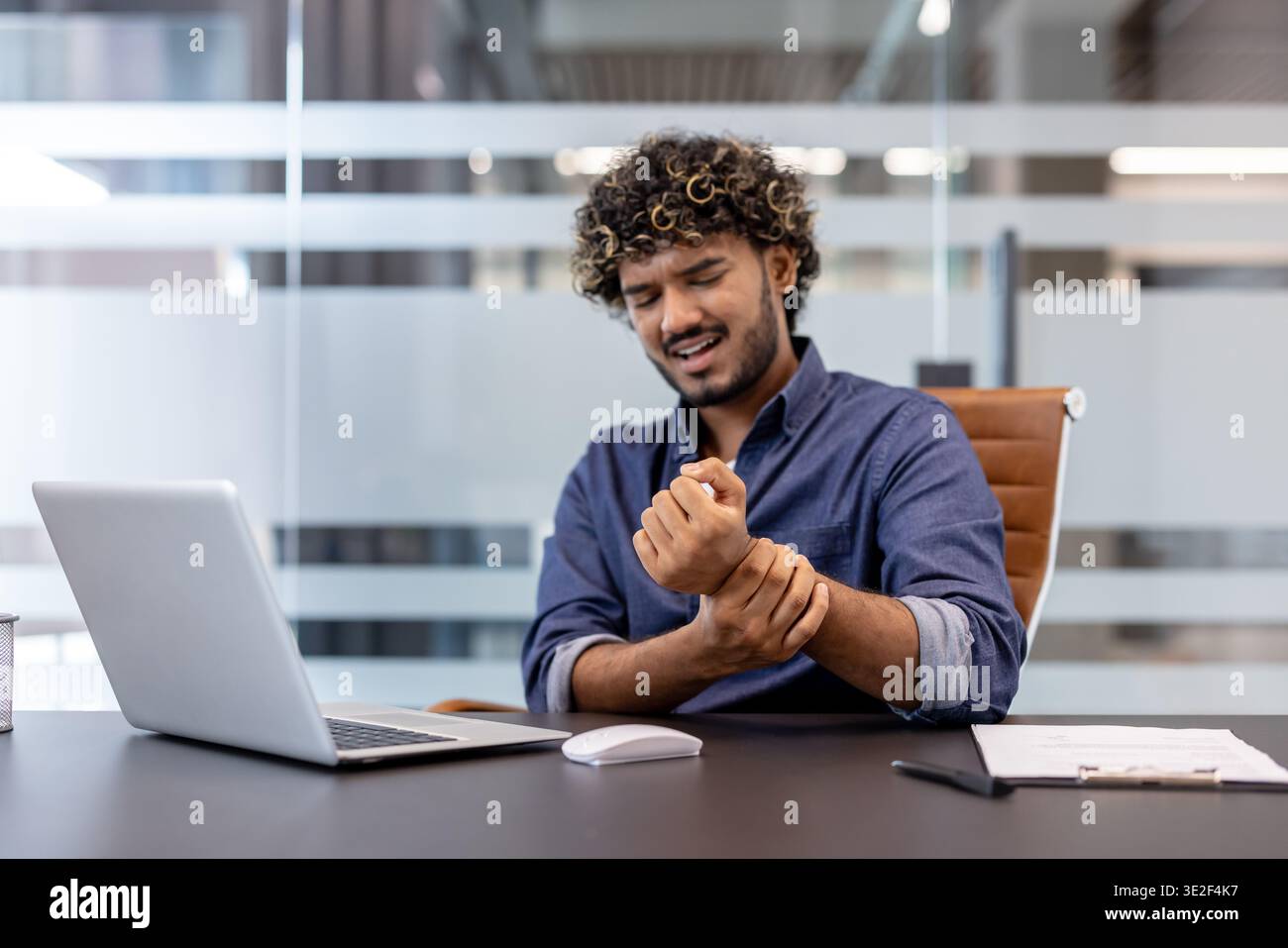Ein junger indischer Mann sitzt an seinem Arbeitsplatz im Büro und hält sein Handgelenk mit der Hand und spürt Schmerzen. Stockfoto