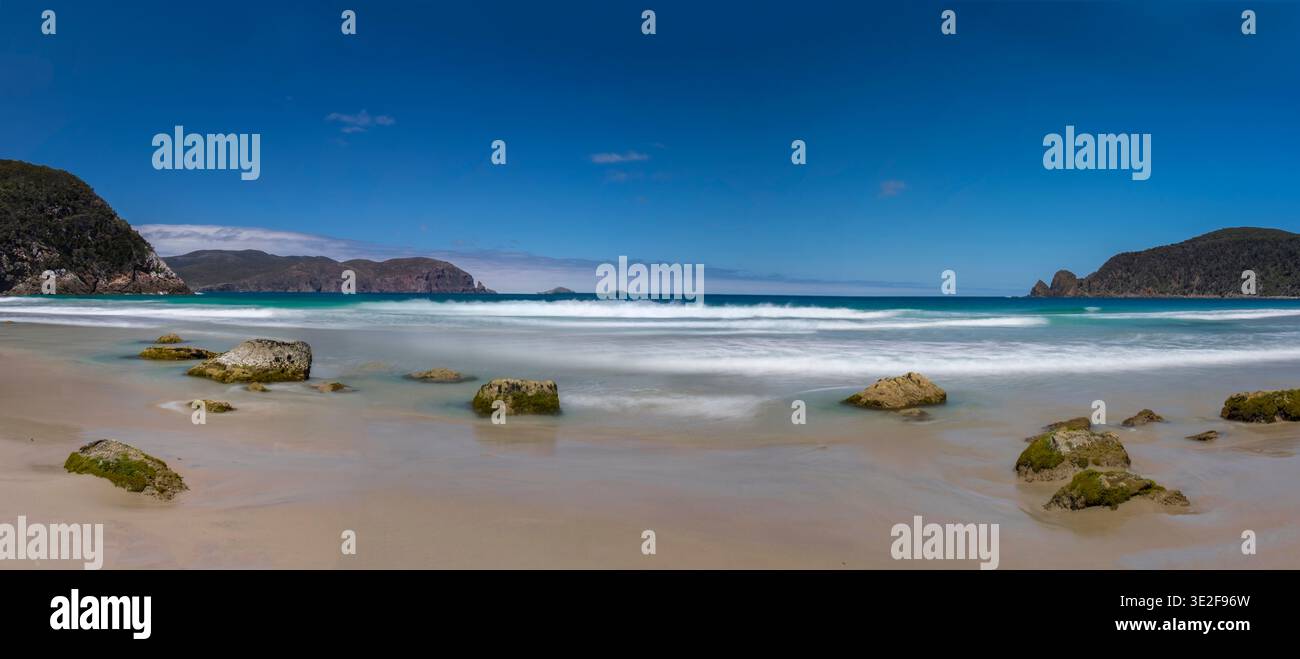 Eine breite tasmanische Strandszene mit Felsen, sanfter Brandung und tiefblauem Himmel. Stockfoto