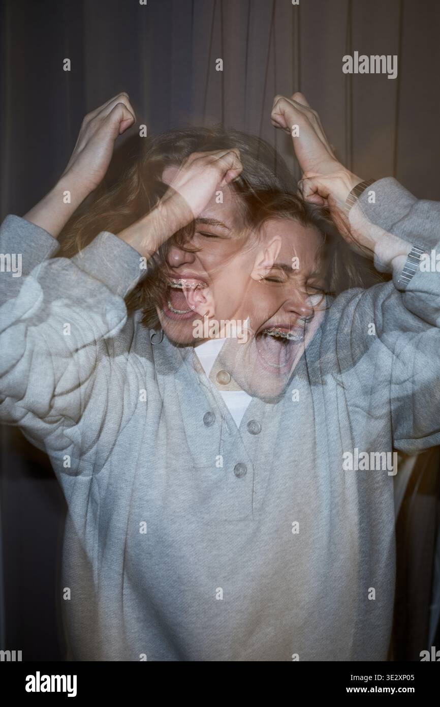 Vertikal der kaukasischen jungen erwachsenen Frau, die intensive Emotionen ausdrückt, Hände hochgestreckt und das Gesicht quälend verzerrt, doppelter Expositionseffekt, der die psychische Belastung betont Stockfoto