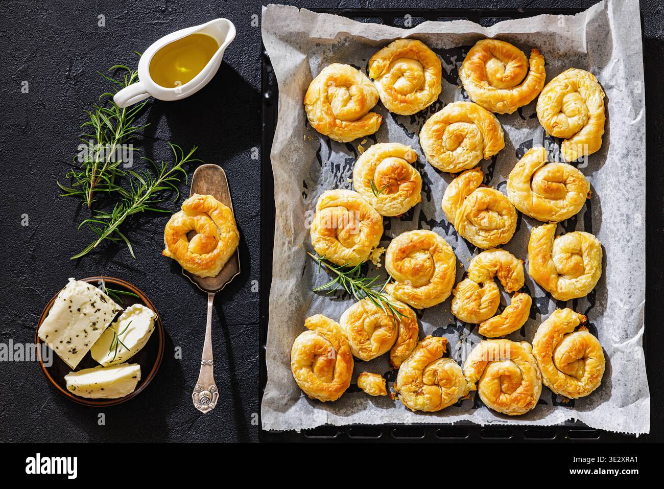 Kreta sarikopita Phyllo Gebäck auf Pergamentpapier in einem schwarzen Backblech auf einem Betontisch mit Rosmarin und Käse, flach gelegt, horizontale Ansicht, cl Stockfoto