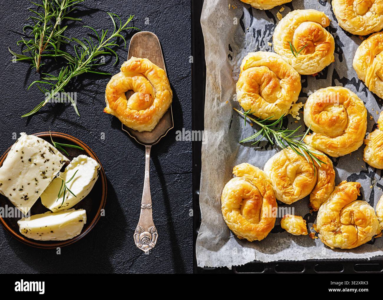 Spiralkäse-Sarikopita-Gebäck auf Pergamentpapier in einem schwarzen Backblech auf einem Betontisch mit Rosmarin und Käse, flach liegend, Nahaufnahme, nicht AI g Stockfoto