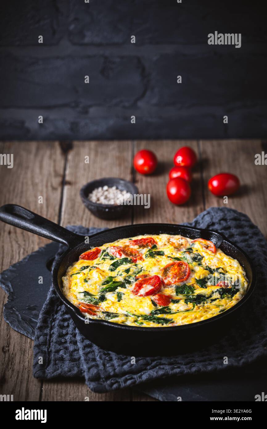 Frittata mit Spinat und Tomate in einer gusseisernen Pfanne auf hölzernem Hintergrund, vertikal mit Kopierraum Stockfoto