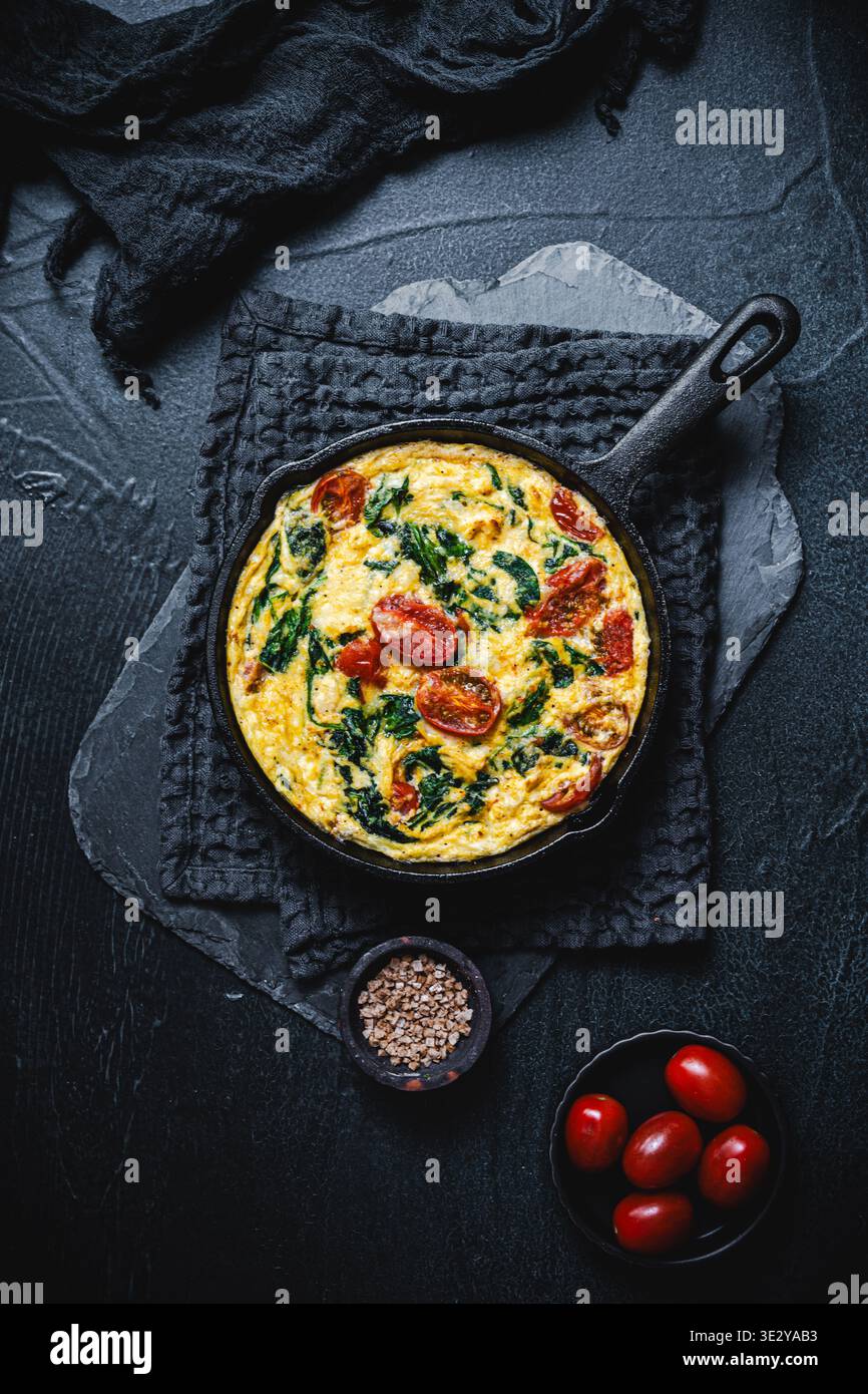 Frittata mit Spinat und Tomate in einer gusseisernen Pfanne auf schwarzem Hintergrund, Draufsicht, Kopierraum Stockfoto
