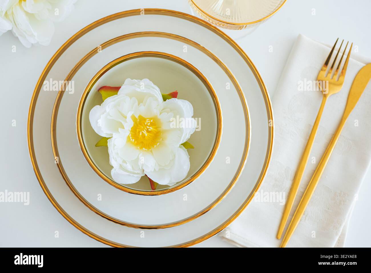 Elegante, universelle Tischeinrichtung mit gestapelten weißen Keramikschalen mit Goldakzenten, einer ordentlich gefalteten Serviette in neutraler Optik und goldenem Besteck Stockfoto