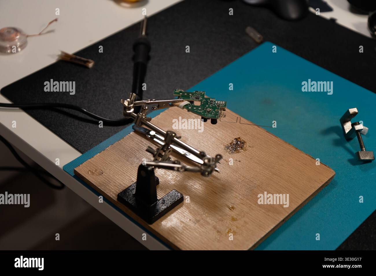 Diese hochwinklige Aufnahme erfasst eine grüne Leiterplatte (PCB), die fest von den Krokodilklemmen eines verstellbaren Lötwerkzeugs mit dritter Hand gefasst wird. Th Stockfoto