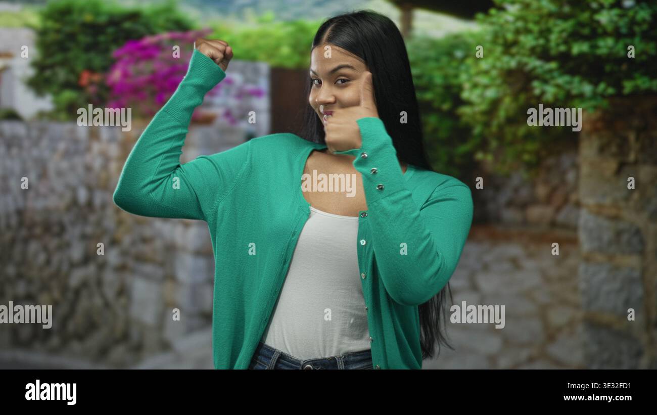 Frau fleht Bizeps in grüner Strickjacke auf einer Steinstraße, die von blühenden Büschen gesäumt ist; Selbstvertrauen stärken Resilienz. Stockfoto