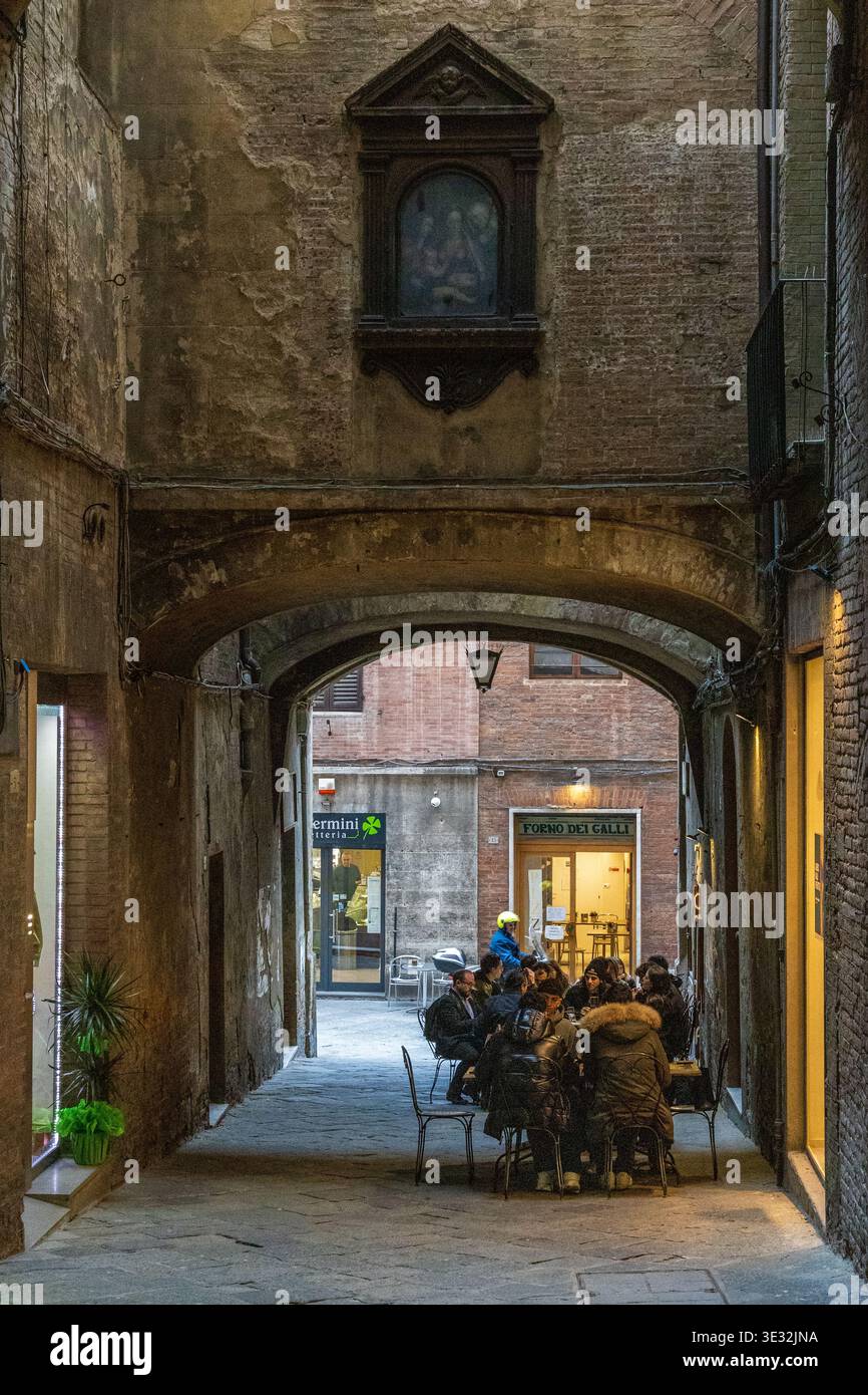 Archway Cafe Siena Im Freien, Abendszene Mit Dinern Unter Warmem Laternenlicht, Schmaler Kopfsteinpflasterallee, Ziegelfassaden Und Intimer Atmosphäre Stockfoto