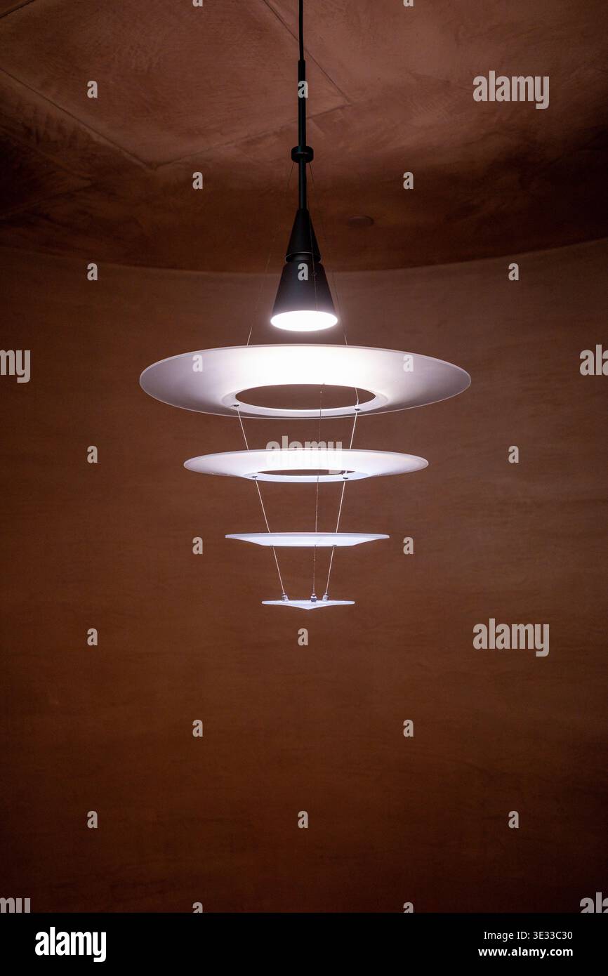 Moderne Hängeleuchte strahlt ein weiches, warmes Licht aus. Kreisförmige Ringe sorgen für einen eleganten, mehrschichtigen Beleuchtungseffekt. Leuchte, die an dunklem Gehäuse aufgehängt ist Stockfoto