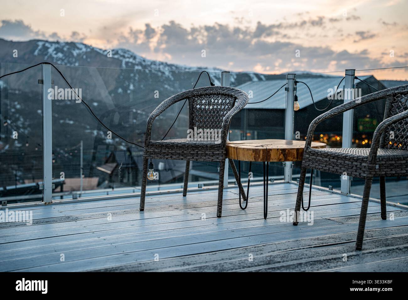 Ein ruhiger Sitzbereich im Freien mit zwei Korbsesseln und einem Holztisch, vor einem Hintergrund der Berge und eines wunderschönen Sonnenuntergangs. Stockfoto
