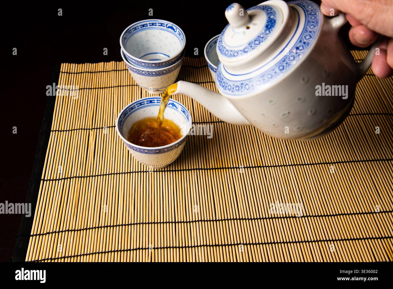 Traditioneller chinesischer Tee, der aus einer Teekanne in eine Tasse gegossen wird, wird auf einer Bambusmatte ausgestellt und hebt die asiatische Teekultur und eine ruhige Präsentation hervor. Stockfoto