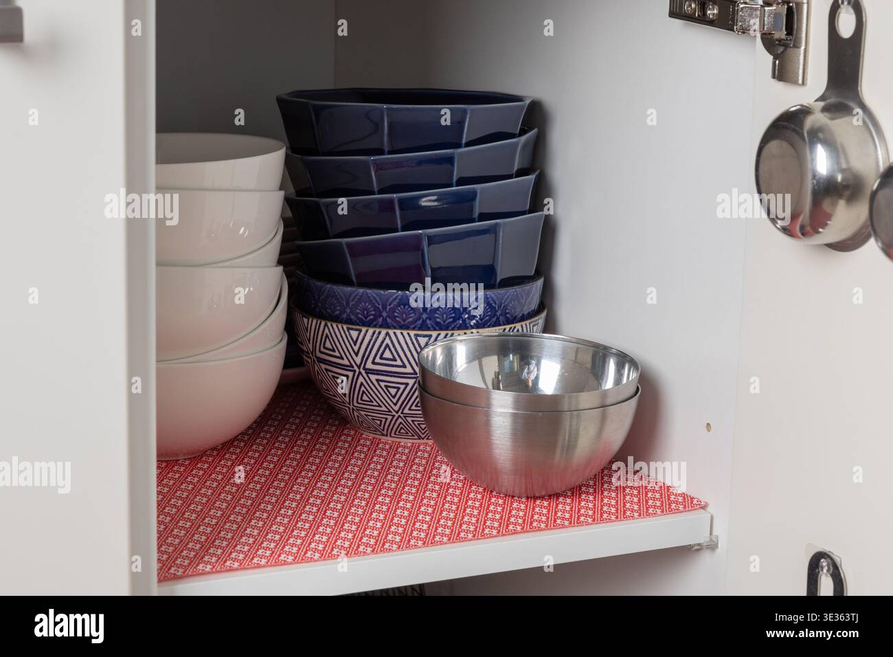 Vorderansicht eines aufgeräumten Küchenschranks mit einer roten Matte, in dem sich stapelbare Keramikschalen und eine Metallschale befinden. Klare Anordnung hebt Ordnung und Einfachheit hervor Stockfoto