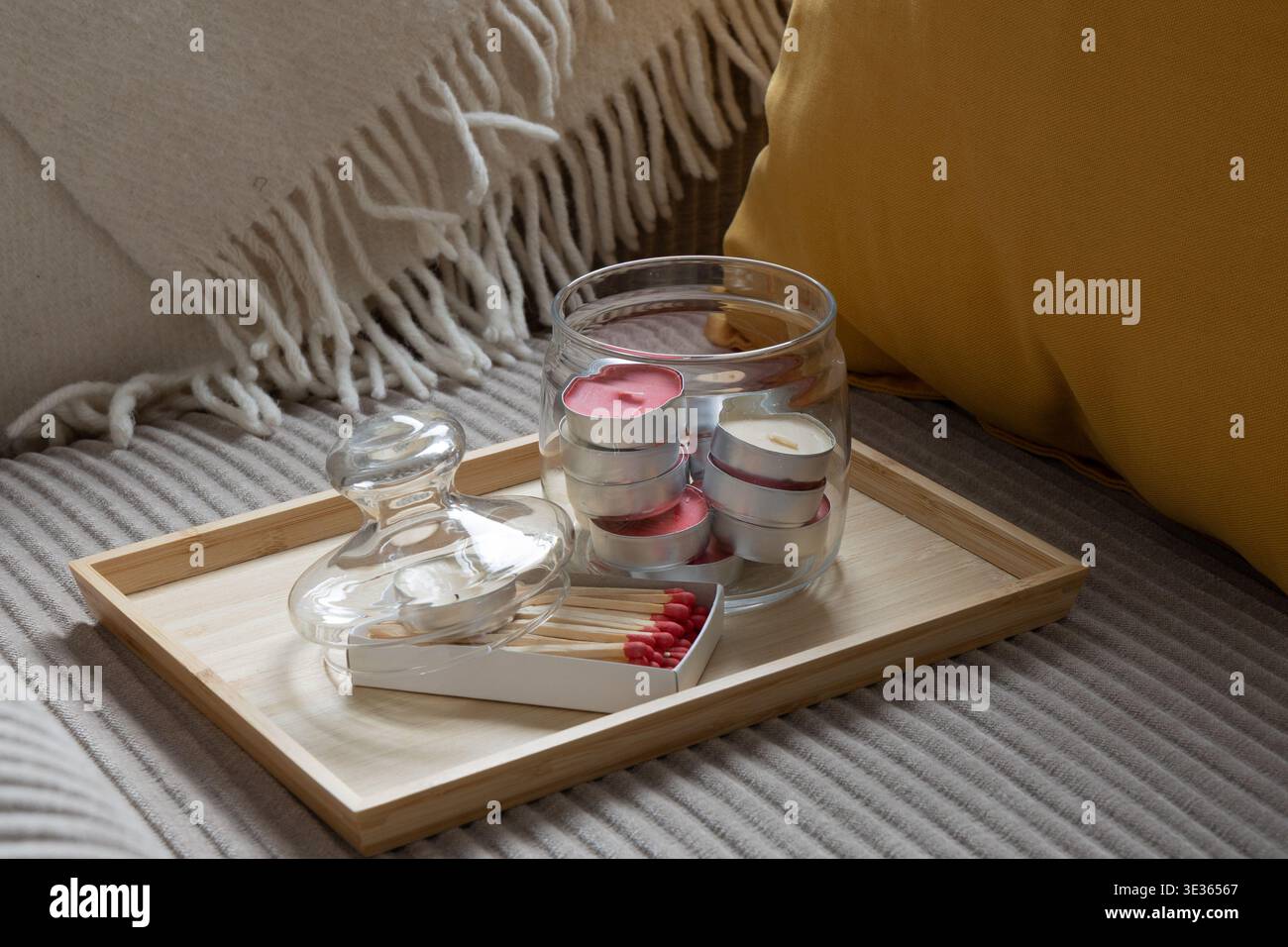 Glasbehälter gefüllt mit Teelichterkerzen und einer Schachtel Streichhölzer auf einem Holztablett über einem weichen Sofa. Warme Textilien und sanftes Licht sorgen für eine ruhige und ruhige Atmosphäre Stockfoto