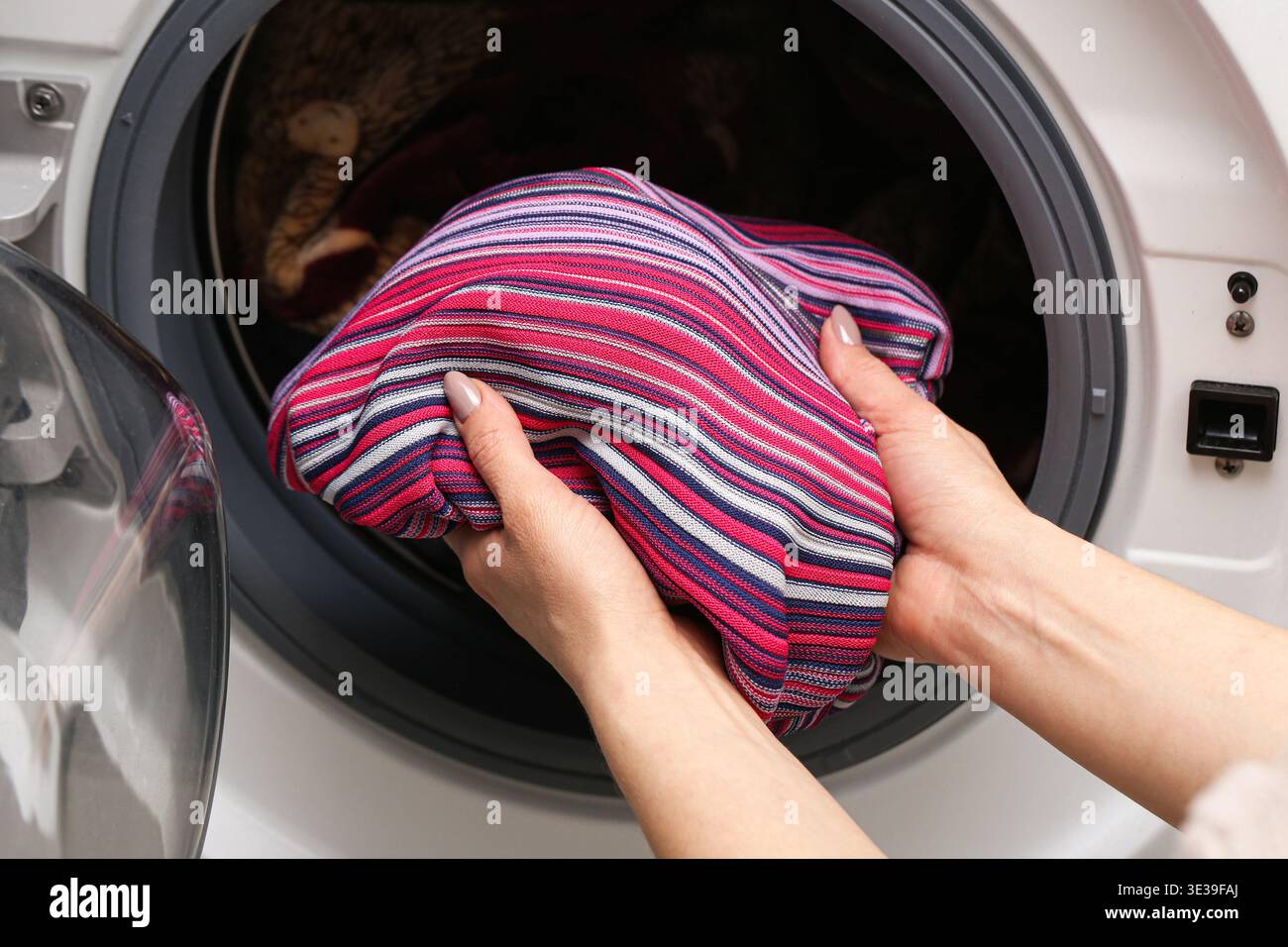 Frau, die bunte gestreifte Kleidung in die Waschmaschine steckt. Wäschetag zu Hause. Hände laden Gewebe in die Waschtrommel. Hausreinigung Routine INT Stockfoto