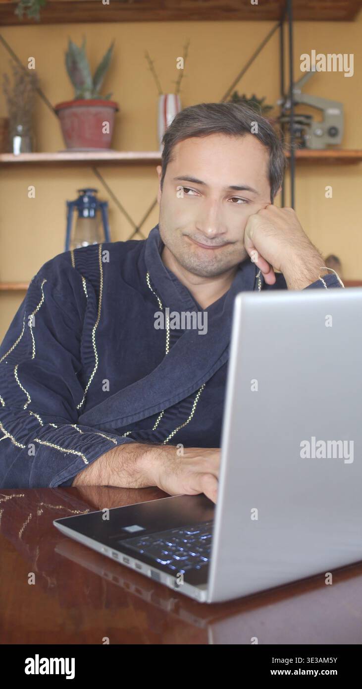 Ein Porträt eines Mannes in einem Morgenmantel, der vor seinem Laptop in Gedanken verloren ist Stockfoto