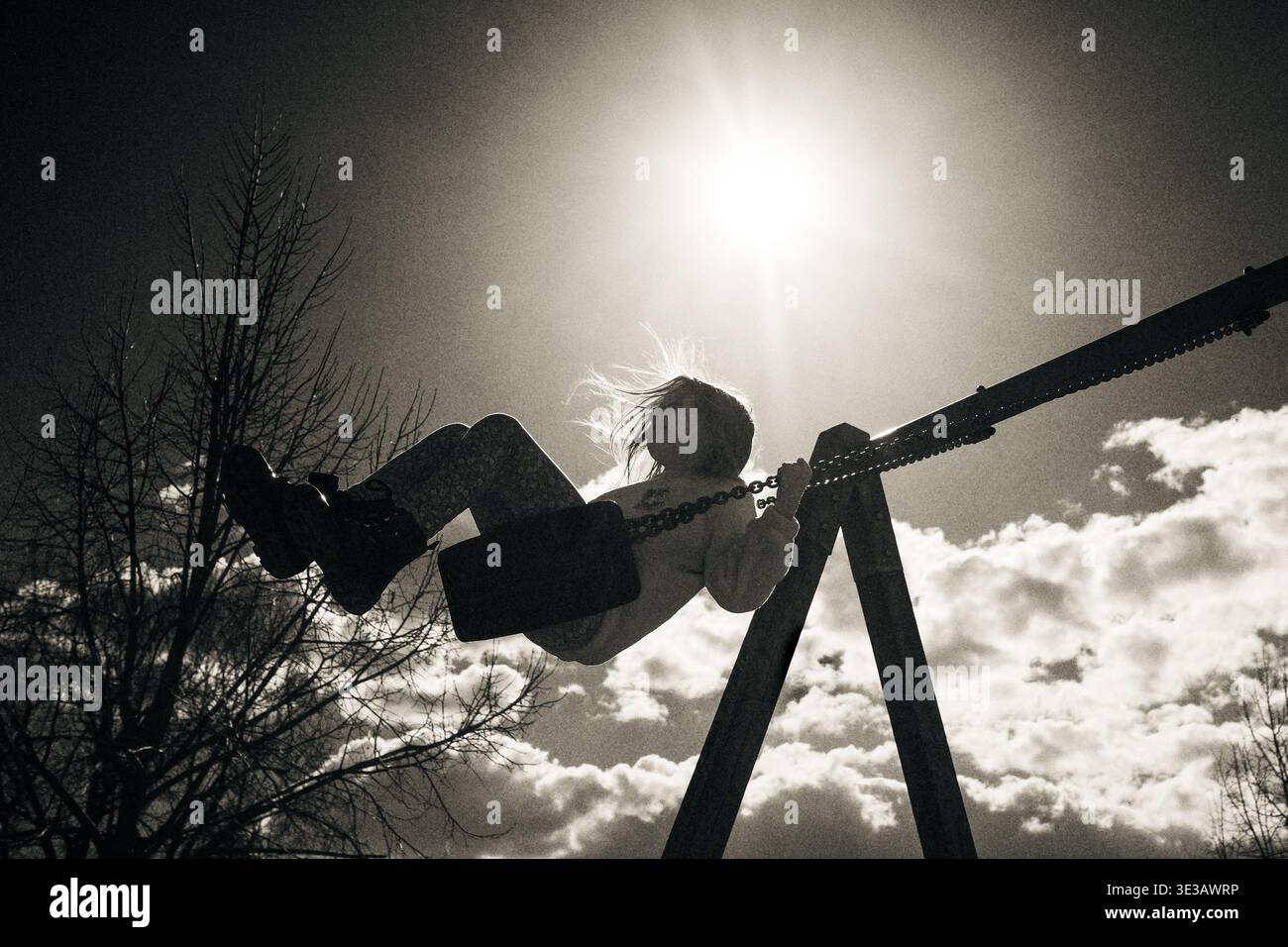 Monochrome Silhouette eines Kindes, das hoch gegen hellen Himmel mit warmem Sonnenlicht schwingt. Emotionale Szene, die Freiheit, Freude und sorglose Kindheit symbolisiert. Stockfoto