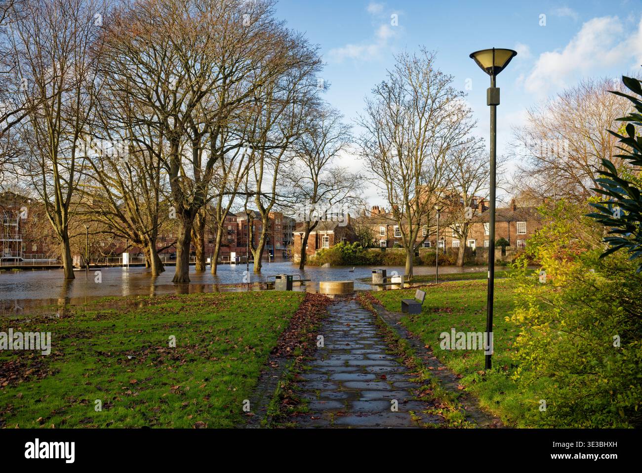 Ein Pfad führt durch einen Stadtpark zu einem überfluteten Gebiet durch einen Fluss mit Häusern im Hintergrund, City of York, England. Stockfoto