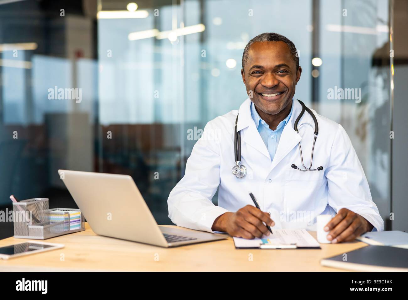 Reifer Arzt im Labormantel und Stethoskop, der selbstbewusst lächelt, während er an einem Schreibtisch mit Laptop und Dokumenten arbeitet und Professionalität und Expertise im Gesundheitswesen verkörpert Stockfoto