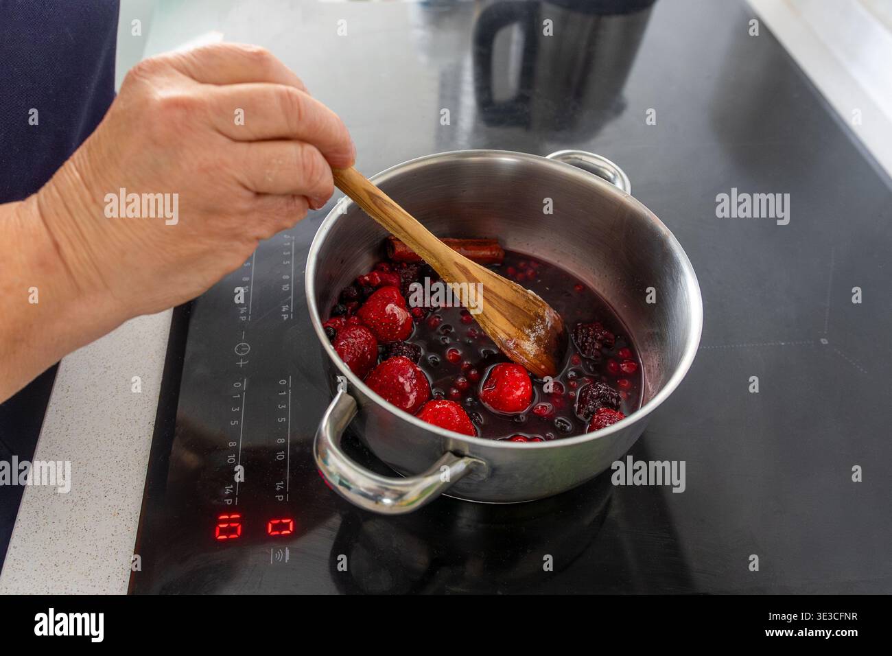 Person, die selbstgemachtes Fruchtkompott oder Marmelade herstellt, indem frische gemischte Beeren in einem Edelstahltopf auf einem Induktionskochfeld umrühren Stockfoto