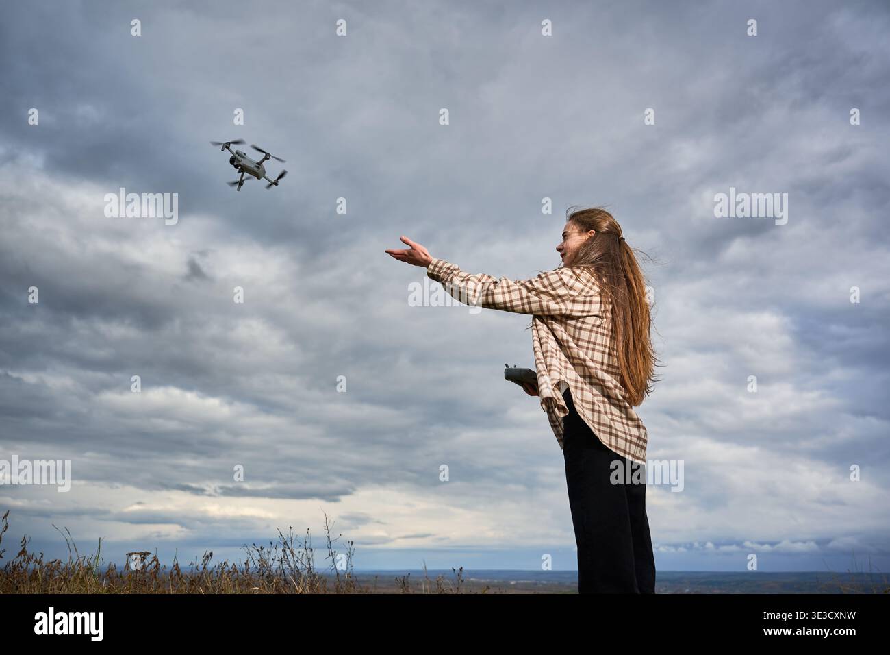 Frau, die in bewölktem Himmel auf fliegende Drohne greift, den Flug mit Fernbedienung auf dem Hügel steuert, weite Sicht auf die Landschaft, Konzept der Technologie, Freiheit und Outdoor-Aktivitäten Stockfoto