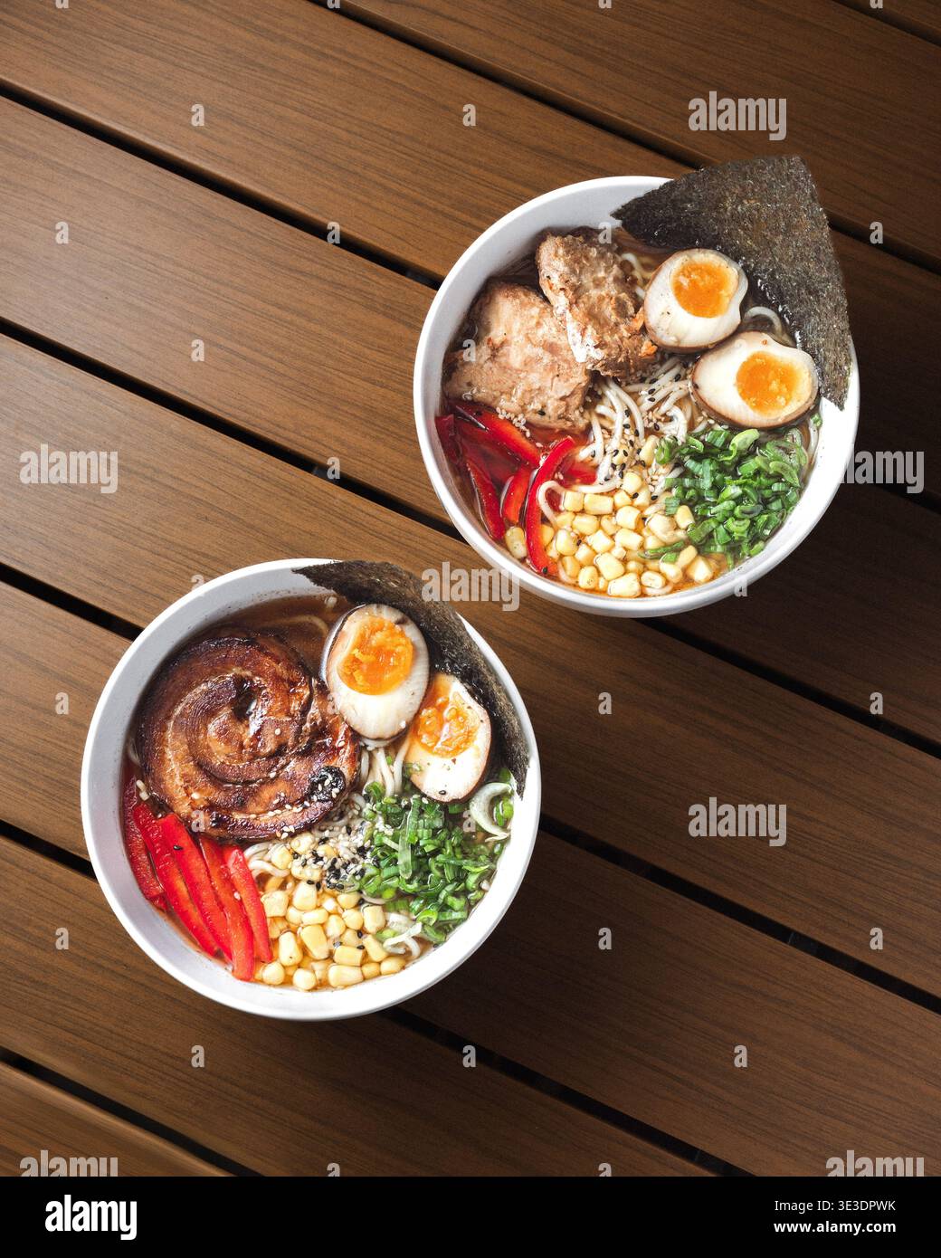 Zwei Schüsseln Ramen ruhen auf einem Holztisch. Die sanfte Beleuchtung hebt die Nudeln und Toppings hervor. Hartgekochte Eier, Gemüse und rote Paprika verleihen Farbe. Tex Stockfoto
