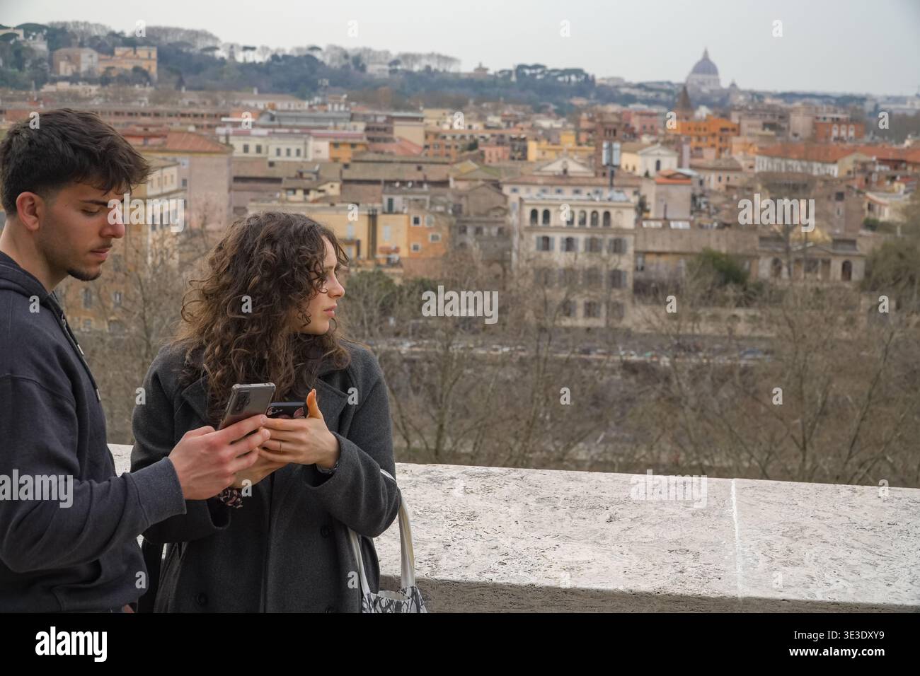 Junges Paar mit Smartphones und Blick auf das weitläufige Stadtbild von Rom, Italien, mit historischen Gebäuden und dem Petersdom in der Ferne. Stockfoto