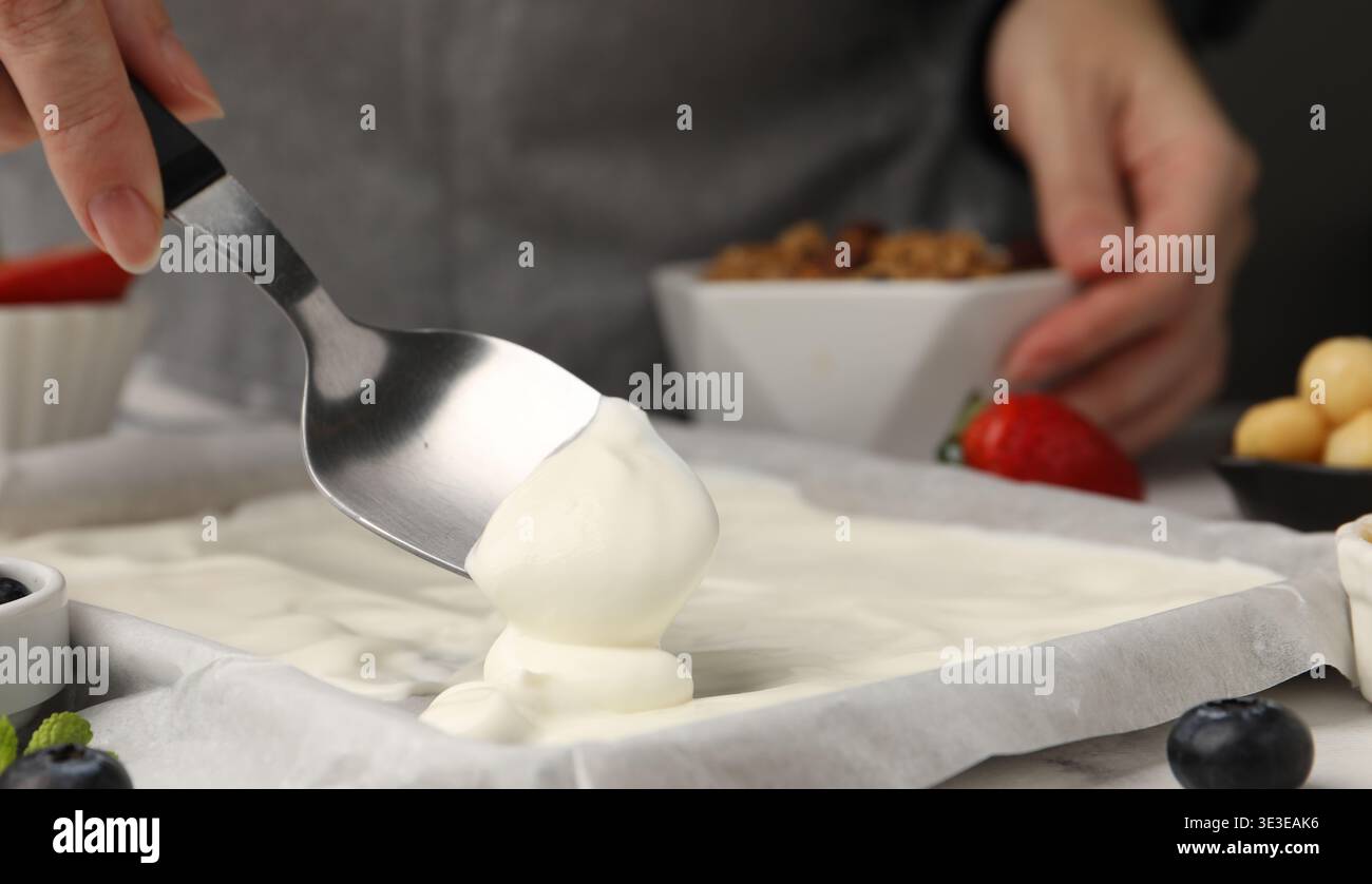 Joghurtrinde herstellen. Frau, die Joghurt mit Löffel auf das Geschirr streut, auf dem weißen Tisch, Nahaufnahme Stockfoto