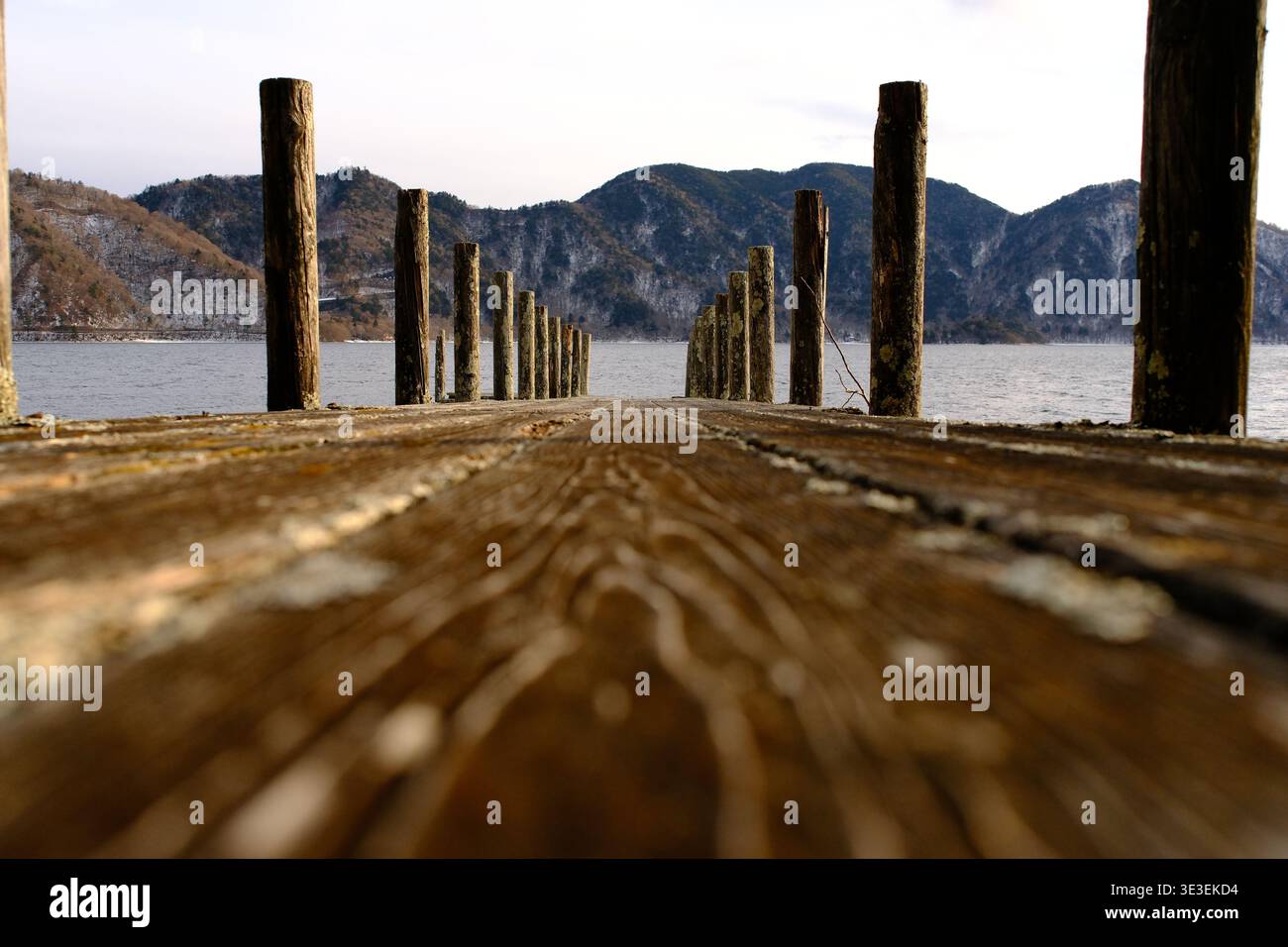 JAPAN – 7. FEBRUAR 2026: Eine Perspektive aus einem flachen Winkel auf einen rustikalen Holzpier, der in Richtung schneebedeckte Berge am Chuzenji-See in Nikko führt. Stockfoto