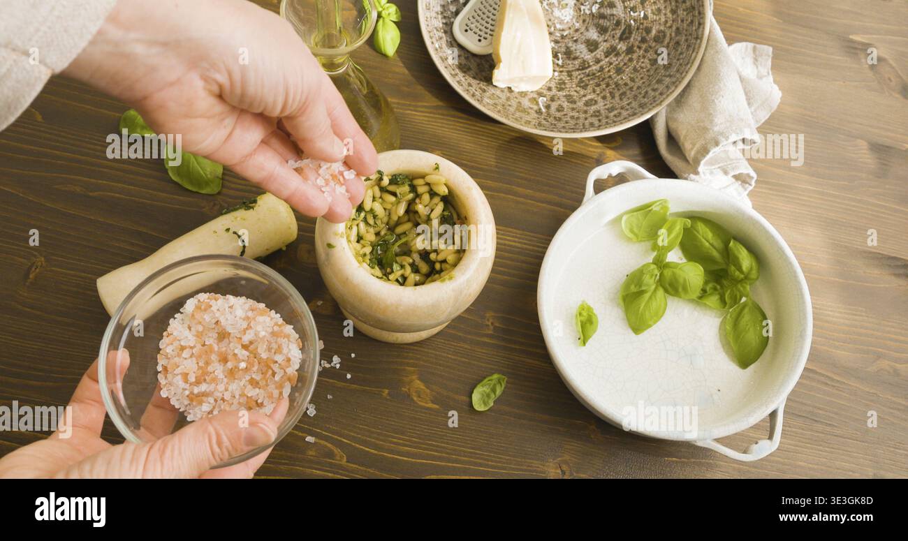 Person fügt rosafarbenes Meersalz in Marmormörtel bei, während Sie traditionelles Genoveses Pesto herstellen. Nahaufnahme der Hände mit einem kleinen Holzlöffel zum Hinzufügen von Himalaya-Salz Stockfoto