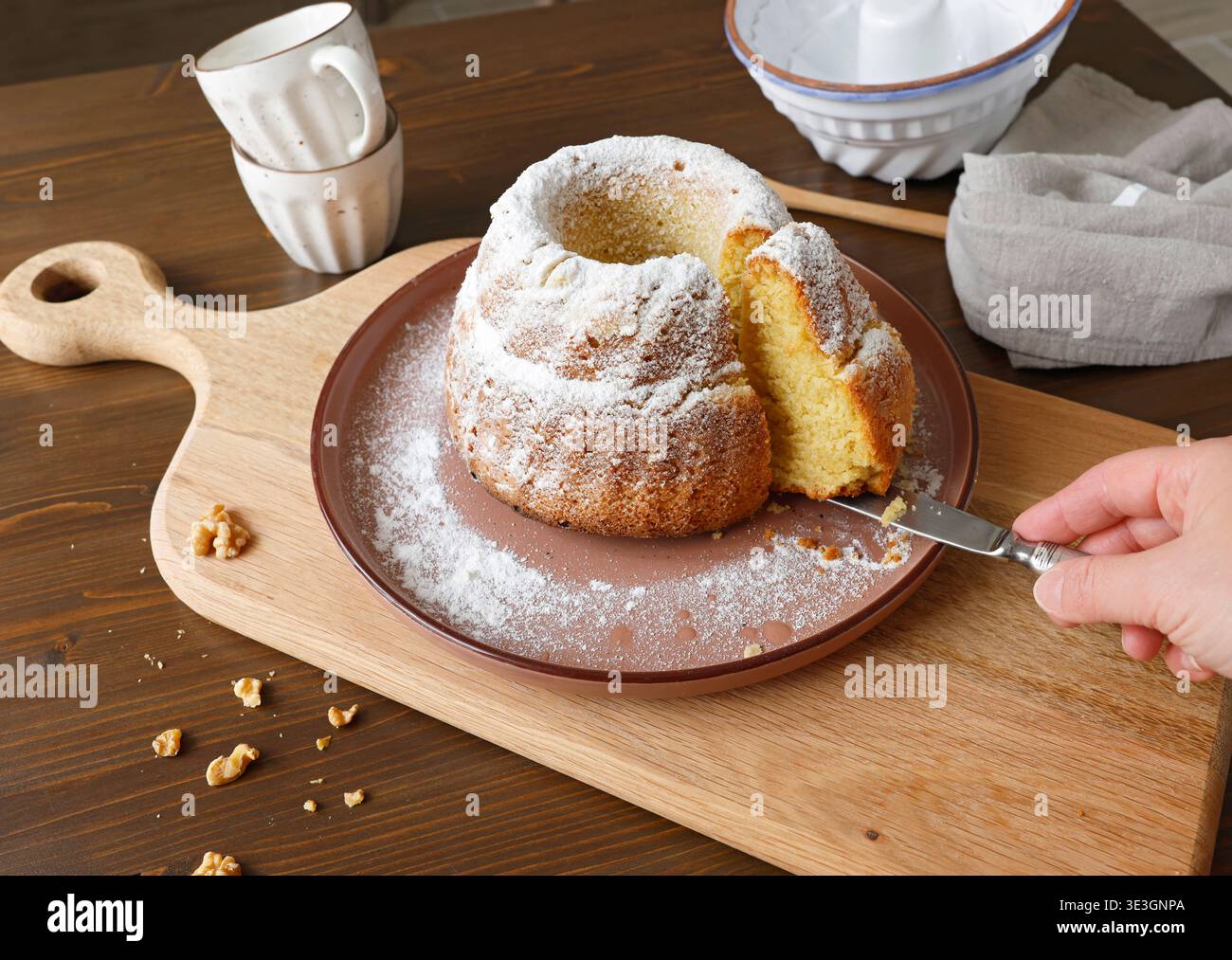Hausgemachter Gugelhupf-Bundkuchen mit Puderzucker auf Holzschneidebrett. Handschneidende Scheibe von frischem Gebäck mit Messer in rustikaler Küche. Stockfoto