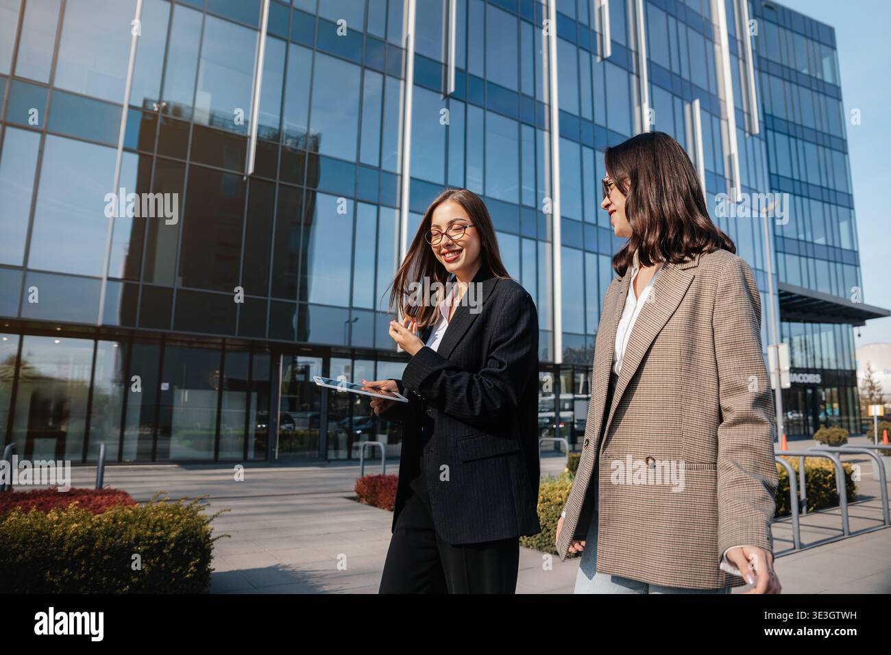 Zwei Frauen laufen zusammen vor einem großen Bürogebäude in der Stadt. Sie sind in Geschäftskleidung gekleidet und führen ein Gespräch, während man hält Stockfoto