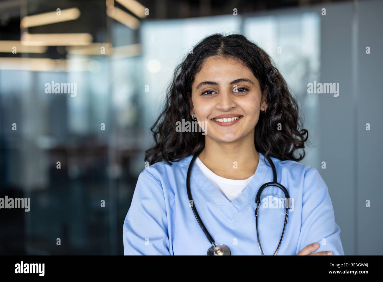 Lächelnde junge Erwachsene indianerin mit hellblauen Peelings und einem Stethoskop, selbstbewusst mit überkreuzten Armen in einer modernen medizinischen Praxis-Umgebung Stockfoto