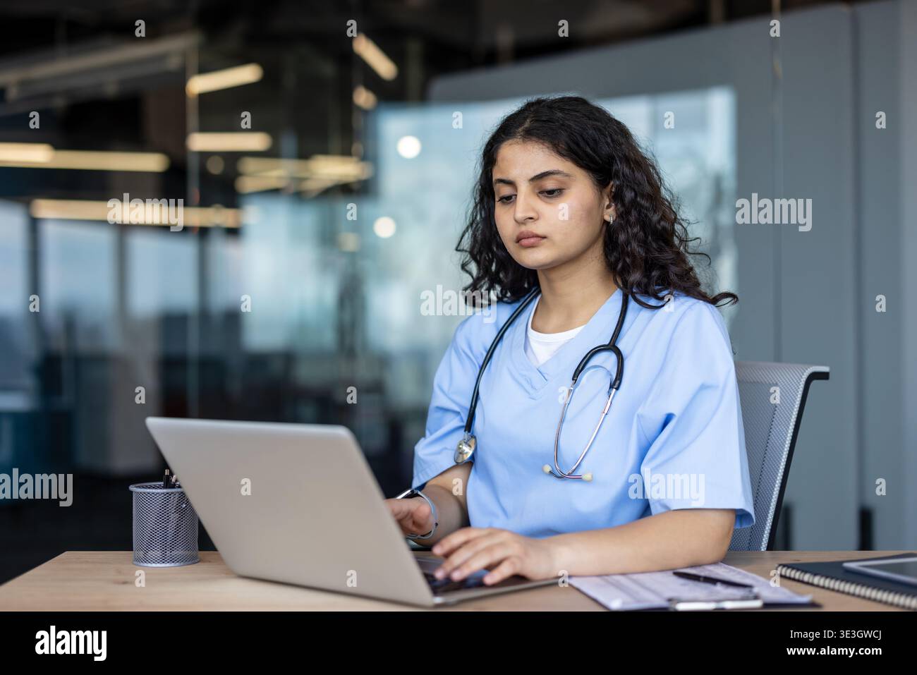 Junge indische Gesundheitsfachkraft mit blauen Peelings und Stethoskop, die sich auf einen Laptop konzentriert, während sie an einem Schreibtisch in einer modernen Klinik oder Krankenhausniederlassung sitzt Stockfoto