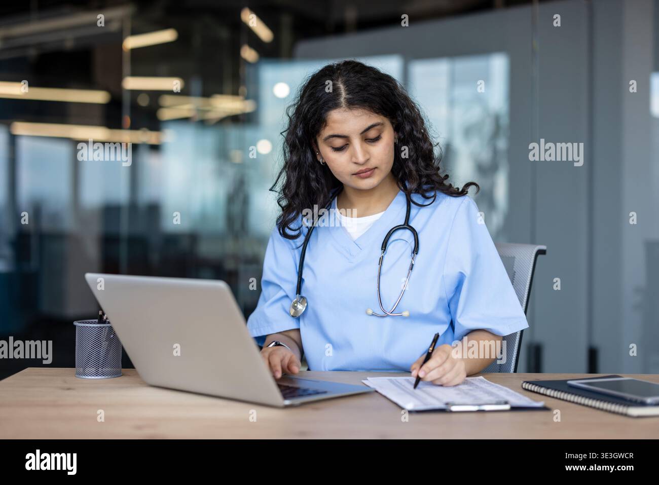 Junge Ärztin oder Krankenschwester in blau schrubbt mit einem Laptop und schreibt Notizen auf eine Zwischenablage am Schreibtisch, wobei der Schwerpunkt auf der Forschung im Gesundheitswesen und der Dateneingabe liegt Stockfoto