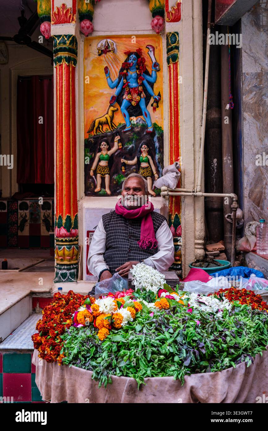 Ein Mann sitzt vor einem farbenfrohen Wandrelief der Göttin Kali, während er im historischen Zentrum von Varanasi Blumenopfer verkauft. Stockfoto