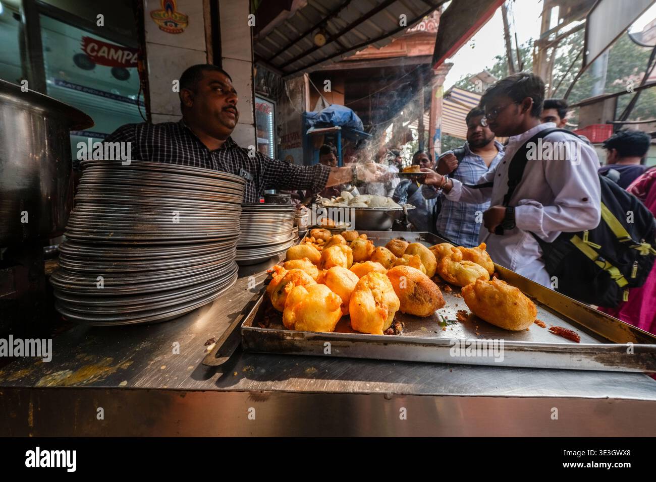 Ein Street Food-Anbieter in Varanasi serviert frittierte Snacks aus einem Metalltablett an Kunden in einem geschäftigen Marktviertel. Stockfoto