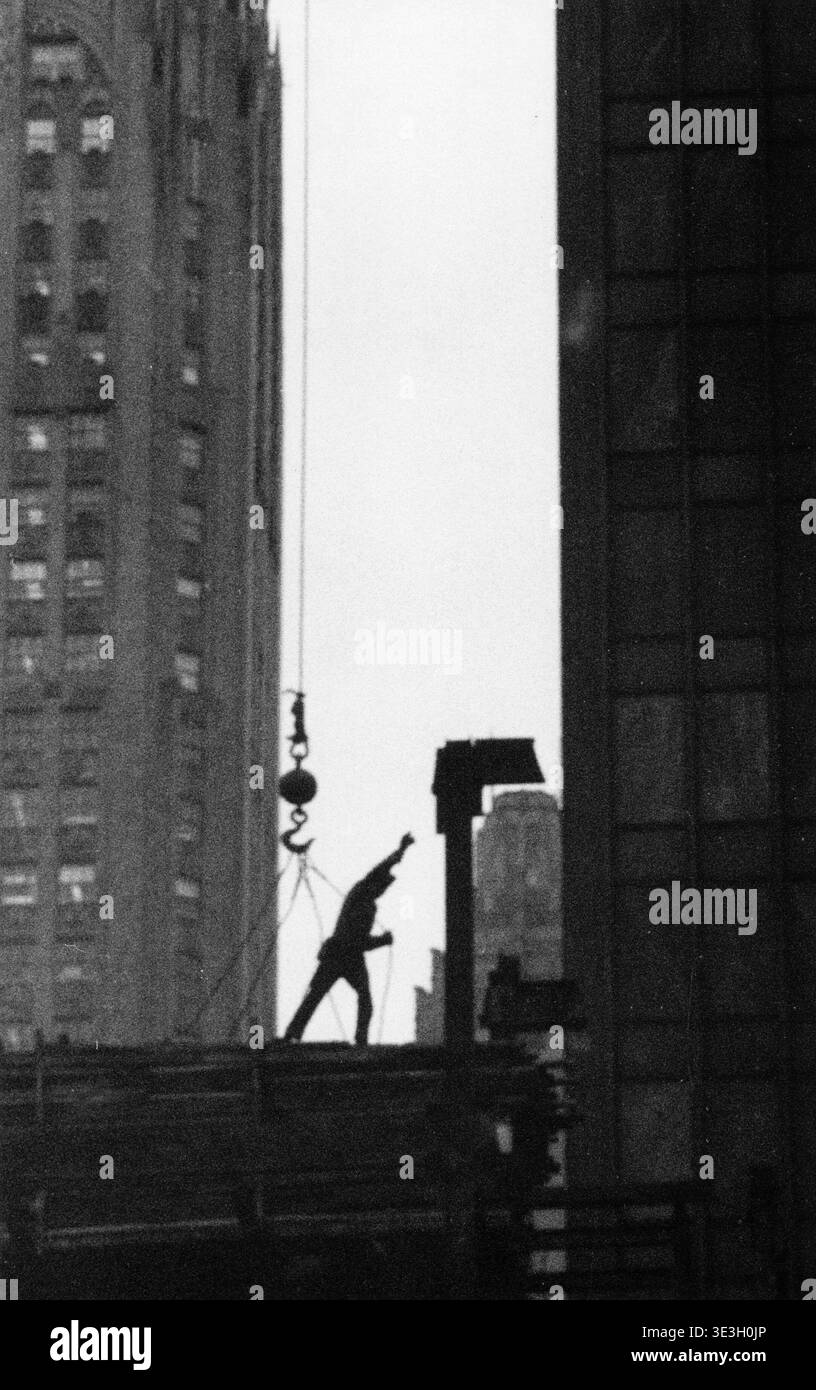 Dieses düstere Straßenfoto, das wahrscheinlich von Angelo Rizzuto in den 1940er Jahren aufgenommen wurde, bildet einen Bauarbeiter inmitten der hohen Wolkenkratzer von New York City dramatisch. Der Arbeiter scheint Baumaterial zu führen, das von einem Kran hochgehoben wird, was das kontinuierliche Wachstum der Stadt und die Arbeit hervorhebt, die mit ihrer beeindruckenden Architektur verbunden ist. Stockfoto