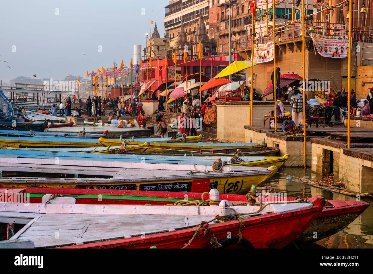 Viele farbenfrohe Holzboote liegen am Dashwamedh Ghat in Varanasi neben dem Fluss Ganges mit traditionellen Regenschirmen und Gebäuden vor. Stockfoto
