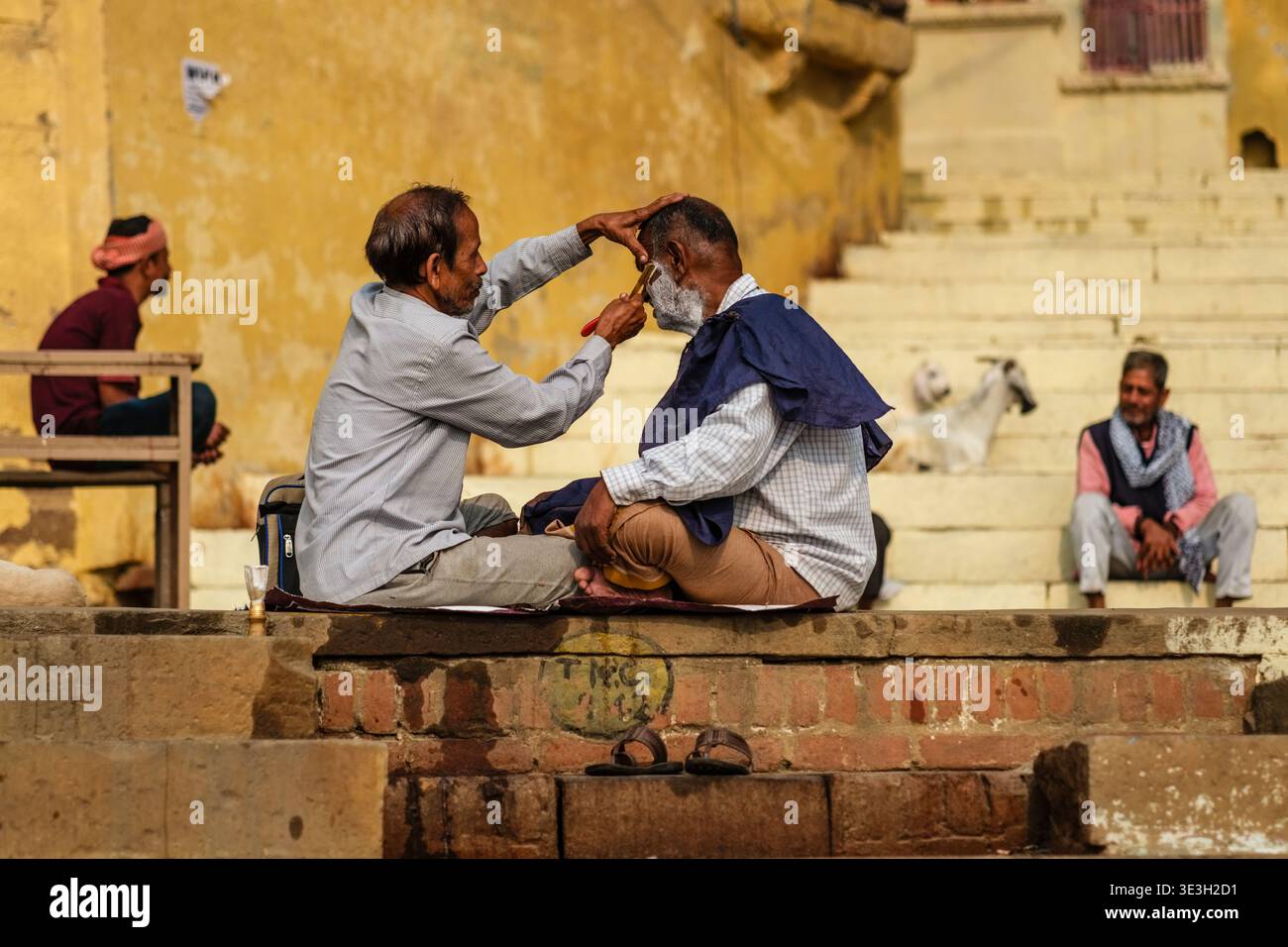 Ein Straßenfriseur rasiert den Bart eines Kunden, während beide im Kreuz auf einem Steinvorsprung in einem Varanasi Ghetto am Ufer des Ganges sitzen. Stockfoto