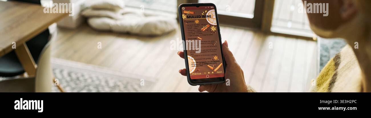 Kopfzeile einer kaukasischen Frau mittleren Alters, die ein Smartphone mit Glühweinrezept hält, drinnen in der Nähe des Fensters sitzt und Sonnenlicht hereinströmt, rechte Greifvorrichtung, partielles Gesicht sichtbar Stockfoto