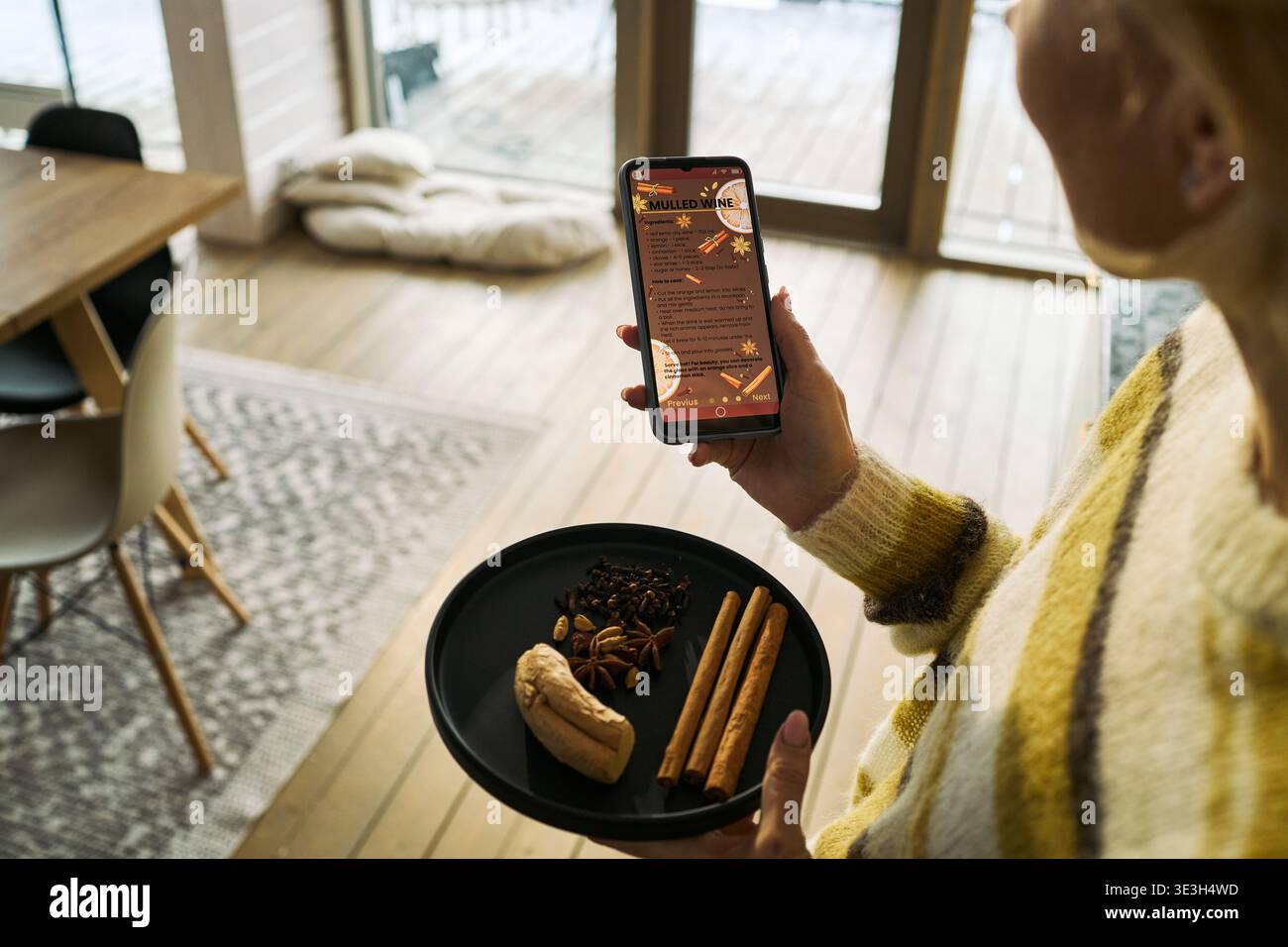 Kaukasische Frau mittleren Alters, die ein Smartphone hält und Glühweinrezept zeigt, während sie einen schwarzen Teller mit Zimtstangen, Sternanis, Nelken und Ingwer in der modernen Küche trägt Stockfoto