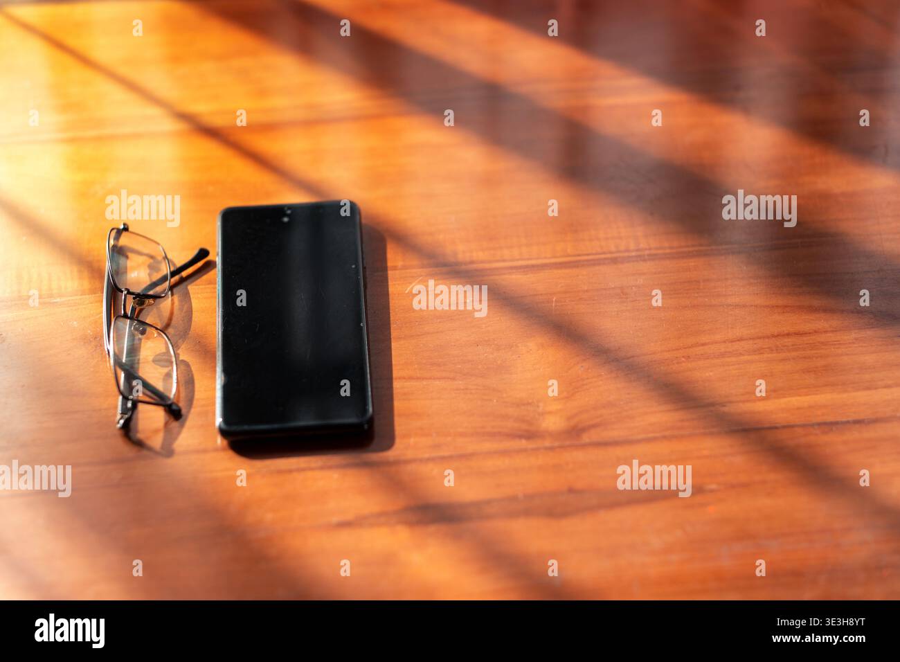 Hochwinkelansicht eines Mobiltelefons und einer Brille auf einer Holzoberfläche, mit langen Schatten aus einem Fenster in warmer Morgensonne. Stockfoto