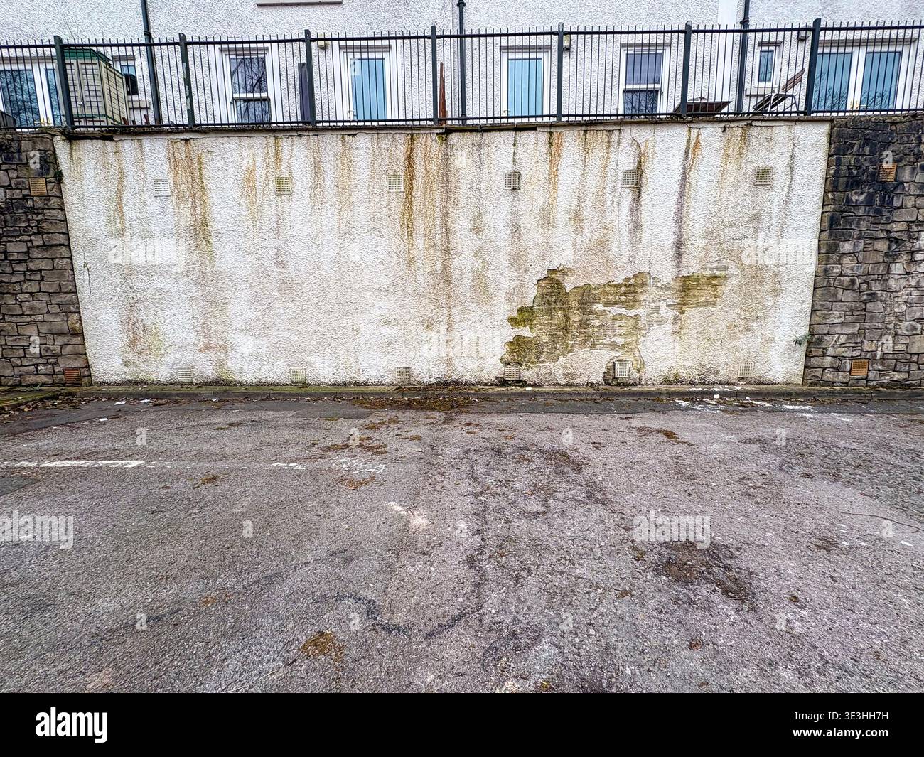 Hinter einem schwarzen Metallzaun ist eine verwitterte weiße Wand mit Roststreifen und Flecken aus grünem Moos zu sehen. Ein paar Fenster und eine kleine grün-weiße Straße Stockfoto