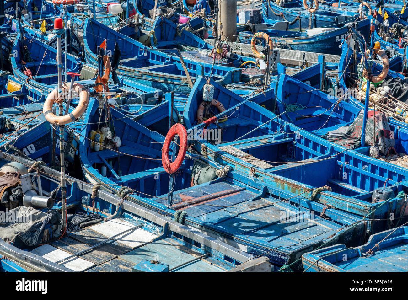 Essaouira, Marokko - Eine dichte Ansammlung von hellblauen Holzfischbooten, die in einem geschäftigen marokkanischen Hafen zusammengebunden sind und Netze, Rettungsringe und Seile zeigen Stockfoto