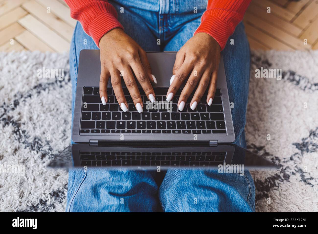 Frau, die einen Laptop auf dem Teppich trägt, während sie zu Hause einen roten Pullover und eine blaue Jeans trägt Stockfoto