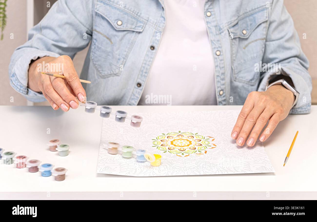 Frau, die Acrylfarbe aus der Palette auswählt, bevor sie das Mandala nach Zahlen malt, kreativer Prozess mit organisiertem Kunstbedarf, Hobby und Kunst Stockfoto