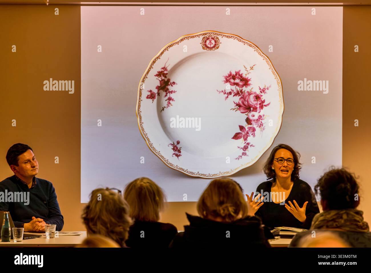 Der Kurator hält einen Vortrag auf einer Porzellanplatte mit violettem Rosendesign und goldenem Rand, die auf die Leinwand projiziert wird. Beachten Sie Hammer und Kompass auf dem Meissener Porzellan. Veranstaltung im Grassi Museum für Angewandte Kunst, Leipzig, Sachsen, Deutschland Stockfoto