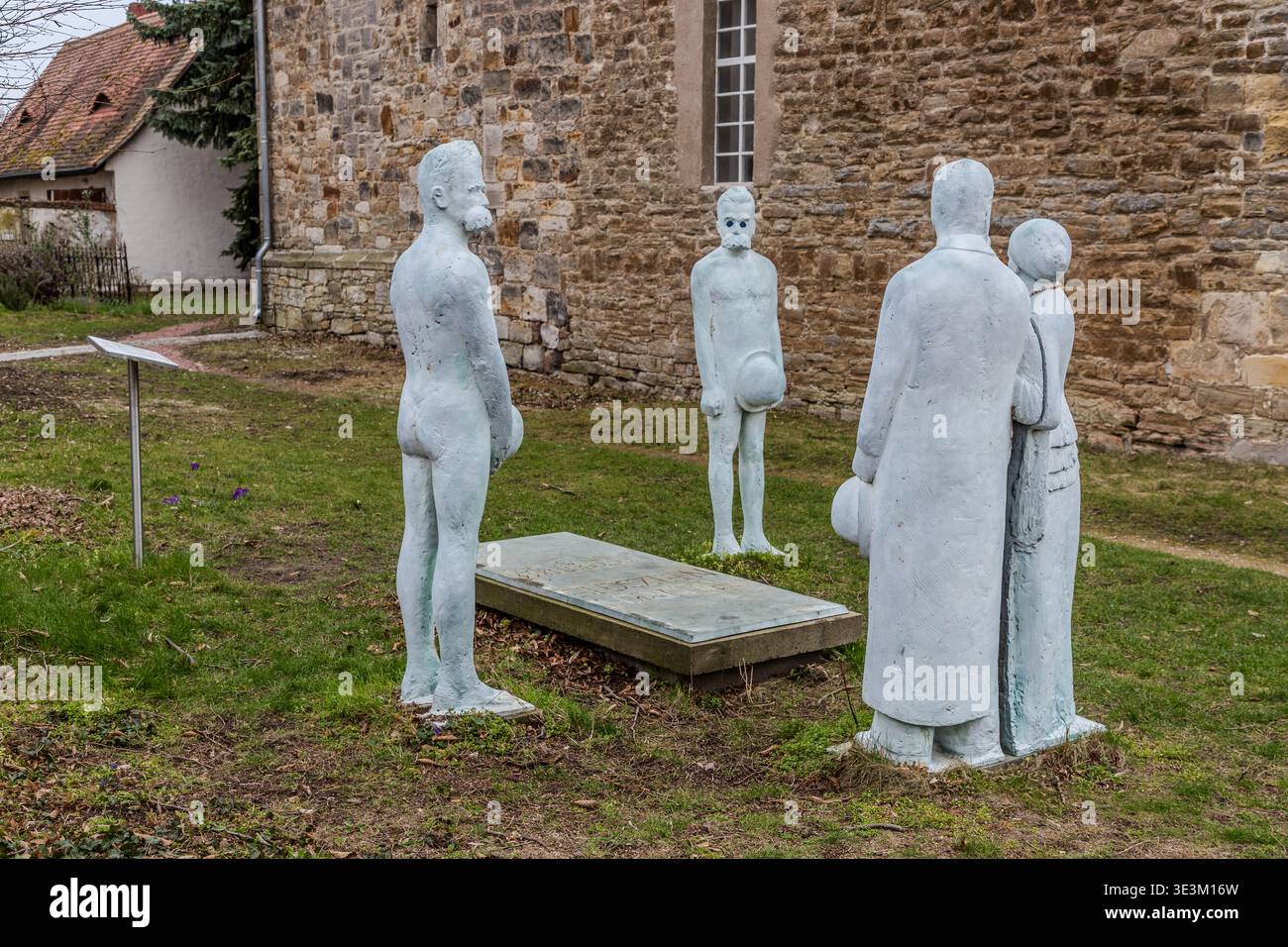 Eine Gruppe von Skulpturen am Grab Friedrich Nietzsches in Röcken: Vier weiße Betonfiguren, die den Grabstein umgeben, mit der Natursteinmauer der Dorfkirche im Hintergrund. Röcken bei Lützen, Sachsen-Anhalt, Deutschland Stockfoto