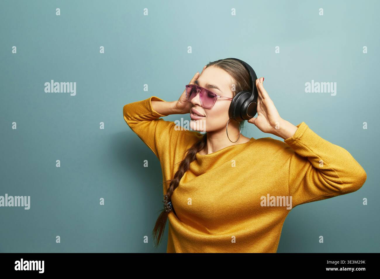 Eine Frau mit langen Haaren hört Musik über Kopfhörer. Sie posiert mit verspieltem Ausdruck, während sie eine rosa Sonnenbrille trägt. Der Hintergrund ist eine einfache Stockfoto