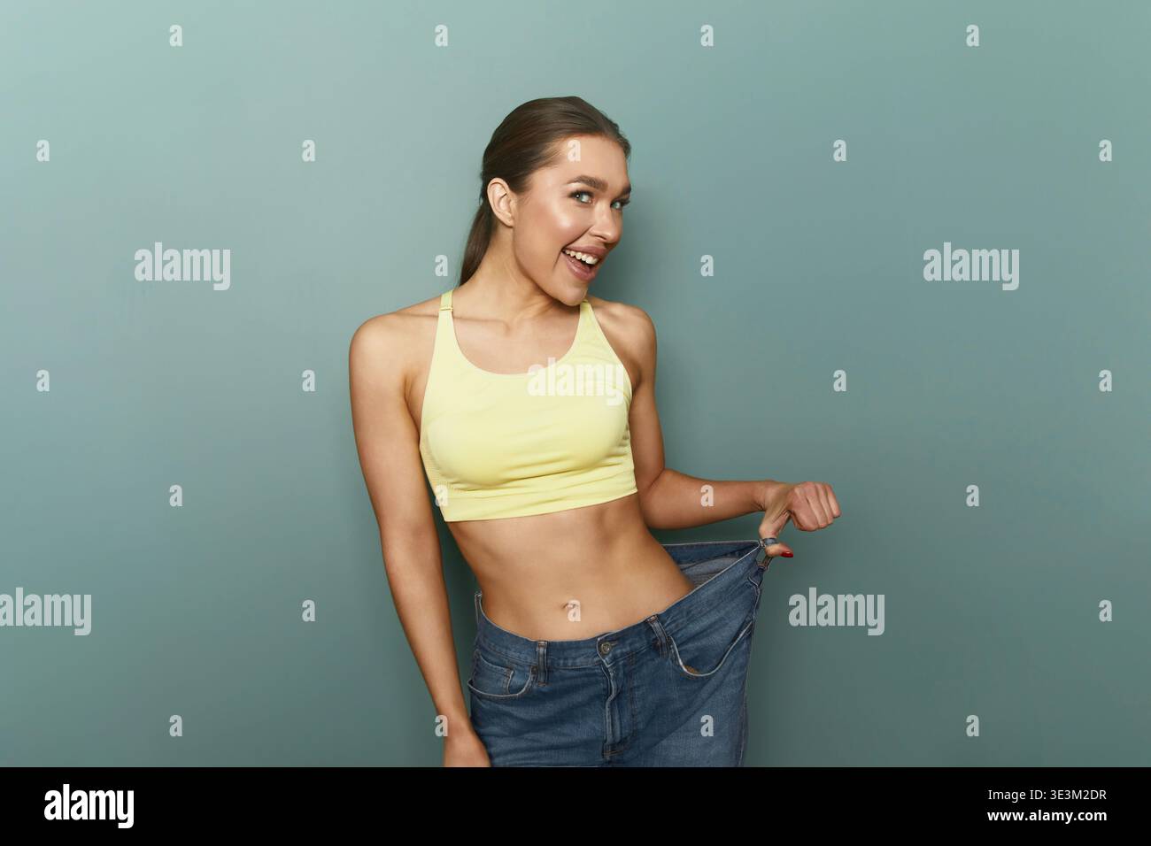Eine Frau steht vor einer grünen Wand und hält ihre lose Jeans mit fröhlichem Ausdruck. Sie trägt ein gelbes Oberteil und scheint die Mo zu genießen Stockfoto